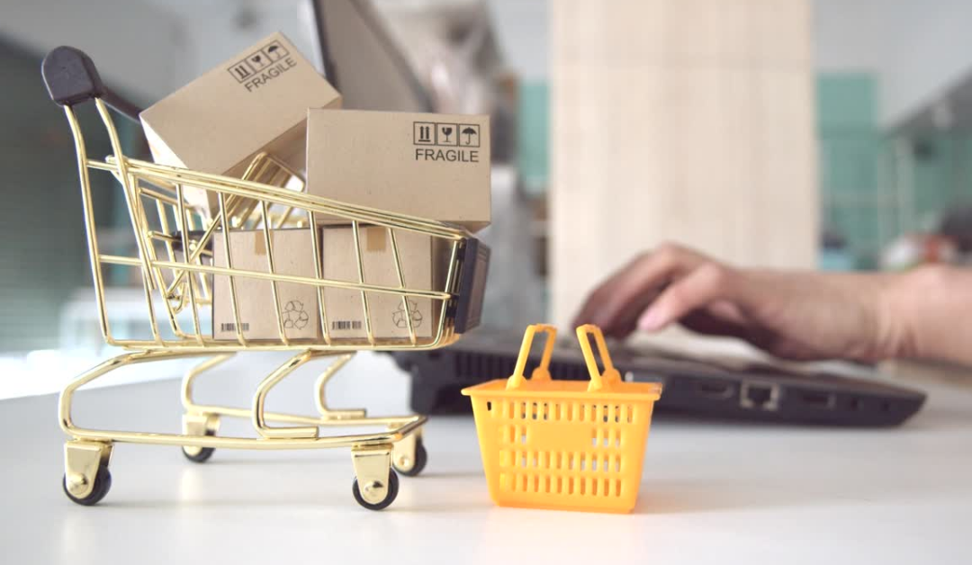 Image of a miniature gold shopping cart with boxes in it labeled fragile sitting on a white counter with a miniature yellow shopping basket sitting on counter to the right of shopping cart, and hands typing on a laptop computer in the background.